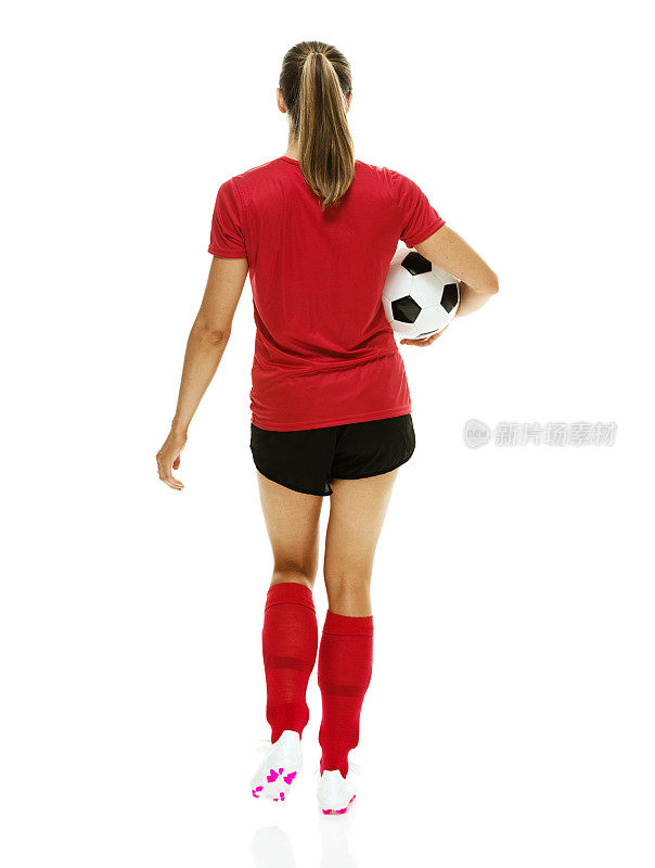 全长/后视图/ 20-29岁的成年人美丽的棕色头发/长发白人女性/年轻女子足球运动员/运动员/女子足球走在白色背景下穿着足球制服和踢足球-运动